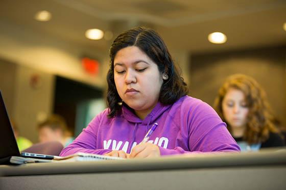 Photo of a Chatham University student writing in a notebook, in a lecture hall. Other students are sitting around her.